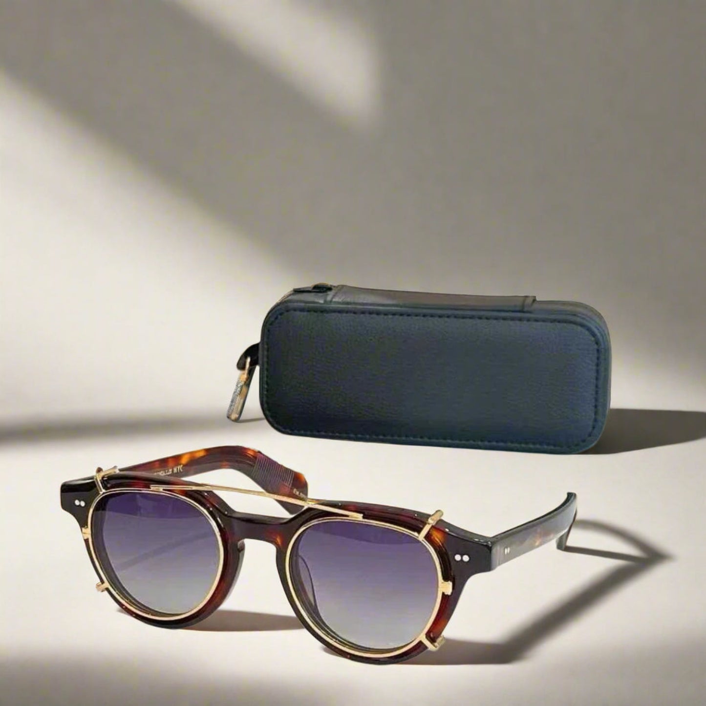 Sunglasses with tortoiseshell frame and purple lenses on a reflective surface with a black case in the background.