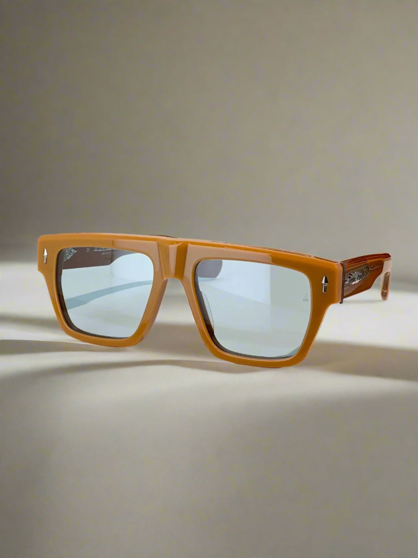 Brown sunglasses with reflective lenses on a wooden stand, with a red box labeled 'Jacques Marie Mige' in the background.