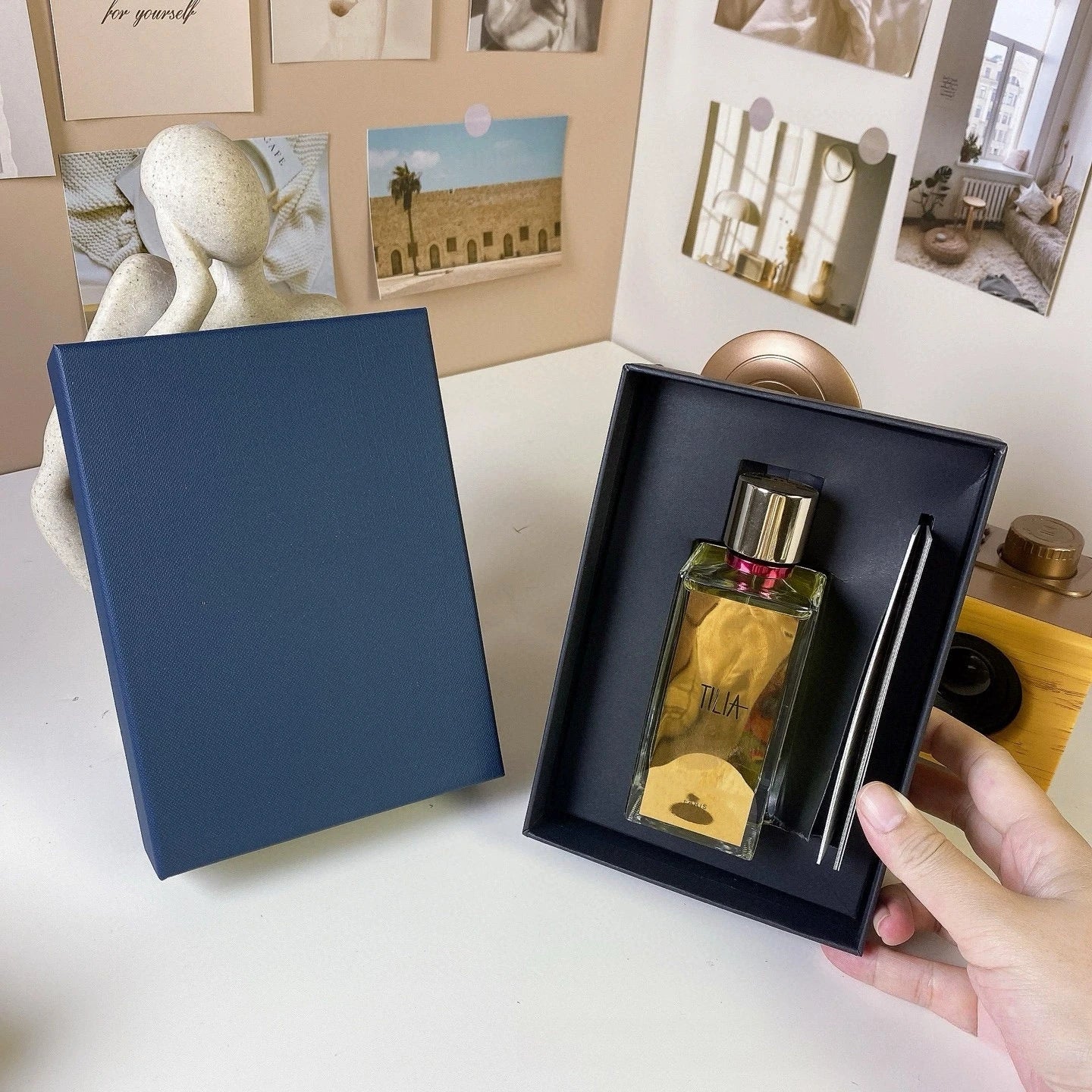 Perfume bottle with a pen in a black box held by a hand, with a blue book and photos in the background.