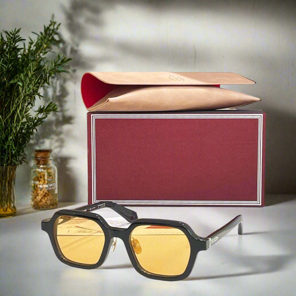 Sunglasses with yellow lenses and black frames on a reflective surface, with a branded box in the background.